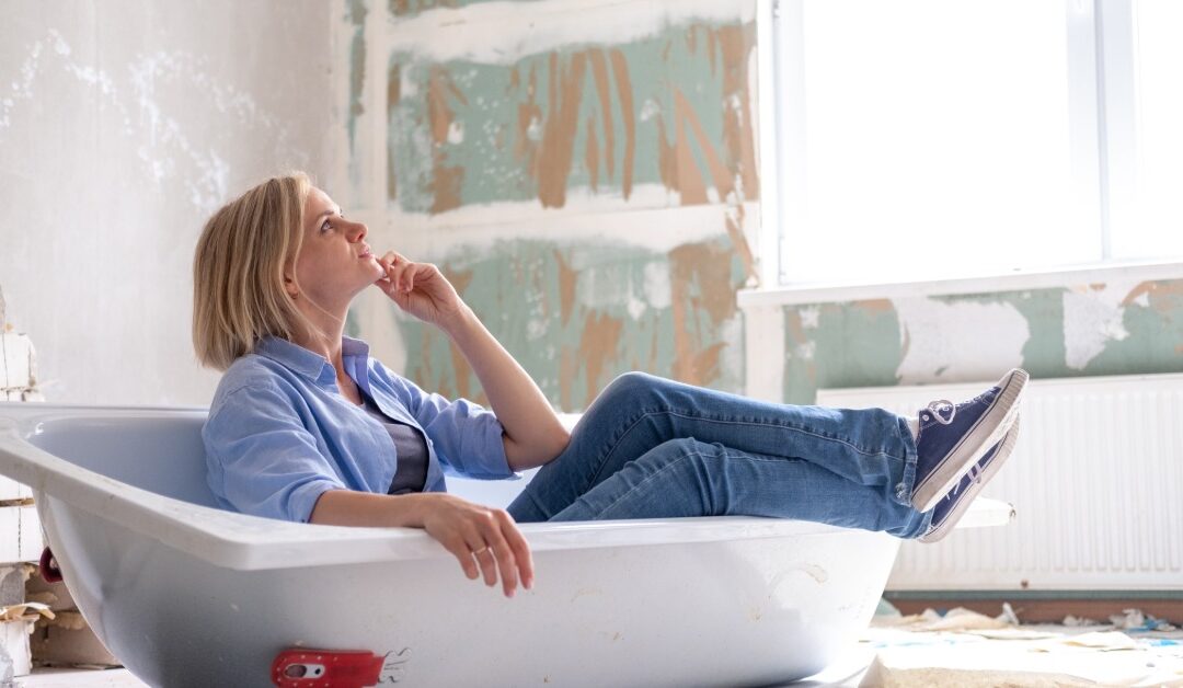 A casually dressed woman looks upward as she sits in an empty bathtub on the ground. The room is partially renovated.