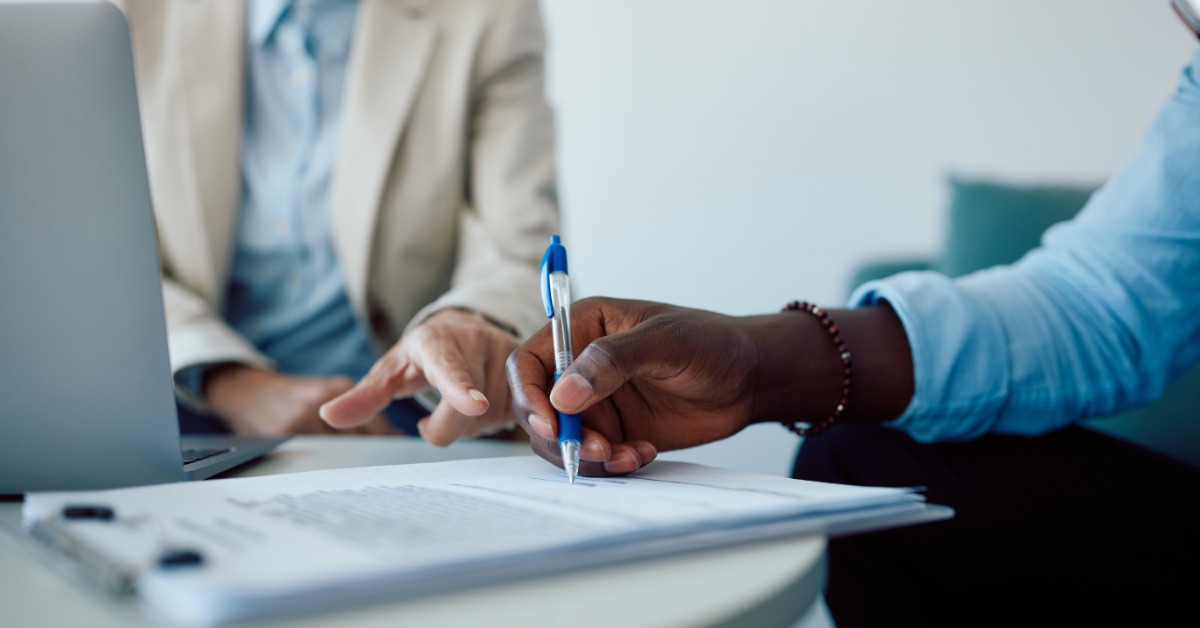 A person wearing a blazer sits behind a laptop and points at nearby paperwork. Another individual signs the document.