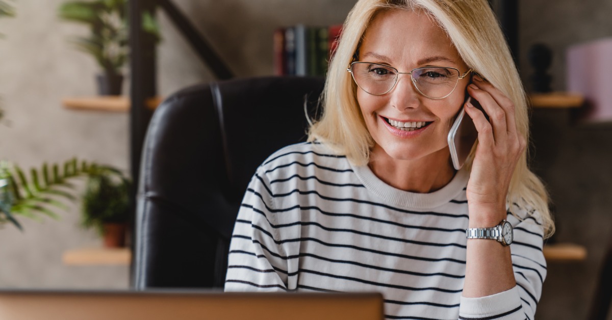 A woman smiles as she talks on a smartphone and looks at her laptop. A bookshelf with books and greenery is behind her.