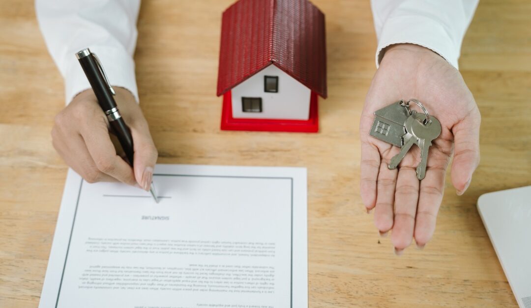 A person holds a set of keys in one hand while signing a document with the other. A small model house is on the table.