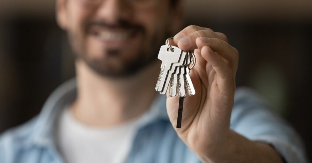 A smiling man holds several keys on a key ring toward the camera. His face is blurred while the keys are in focus.