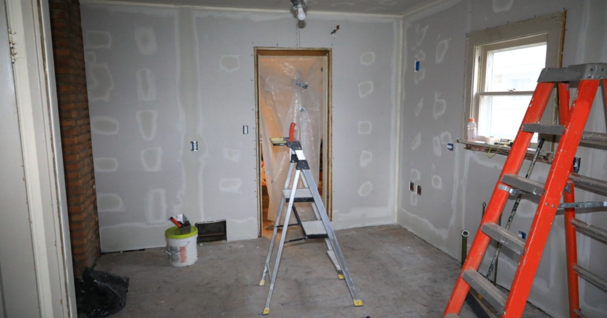 Interior room under renovation with unfinished drywall, visible joint compound spots, a concrete floor, and two ladders.