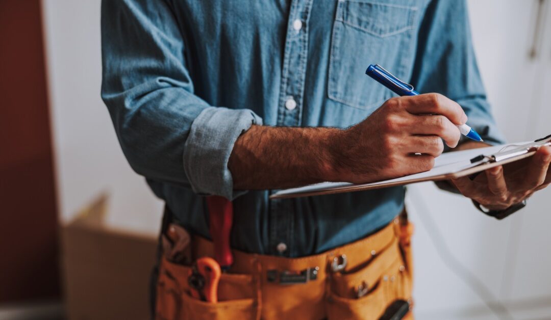 Close-up of a person in a denim shirt holding a clipboard and writing with a pen. They wear a tan tool belt with tools.