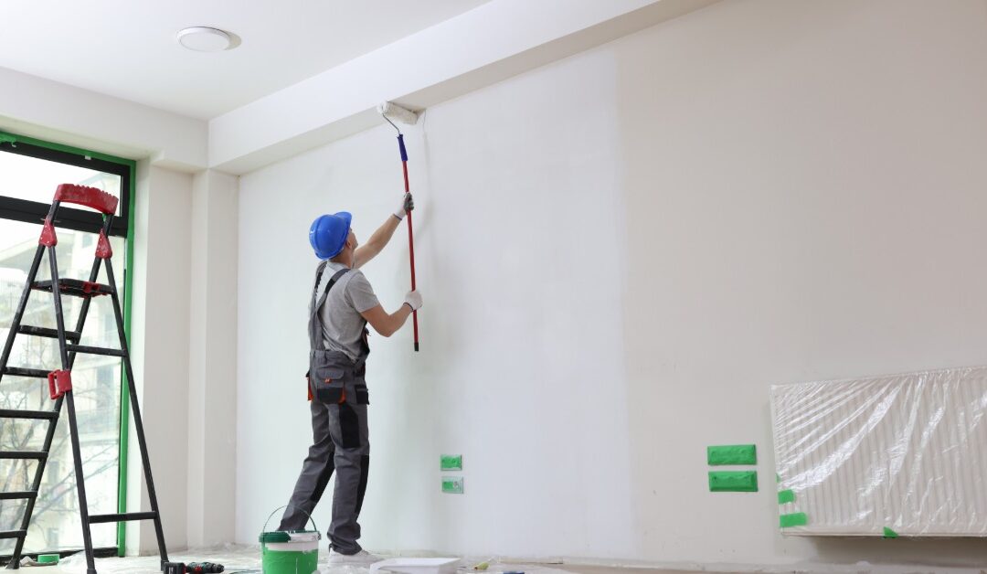 A worker in a hard hat uses a long-handled roller to paint a white interior wall. His ladder is near a large window.