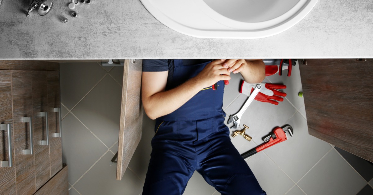 Top-down view of a countertop and sink basin. A person in coveralls works under the sink with tools spread nearby.