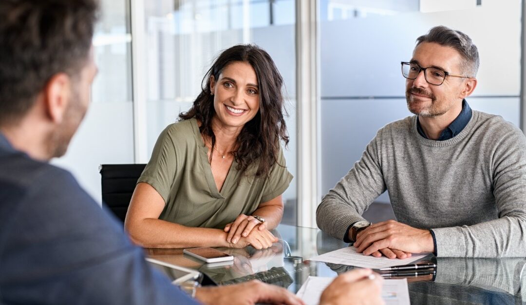 A man and a woman sit side by side at a conference table facing another person. Papers and a tablet rest on the table.