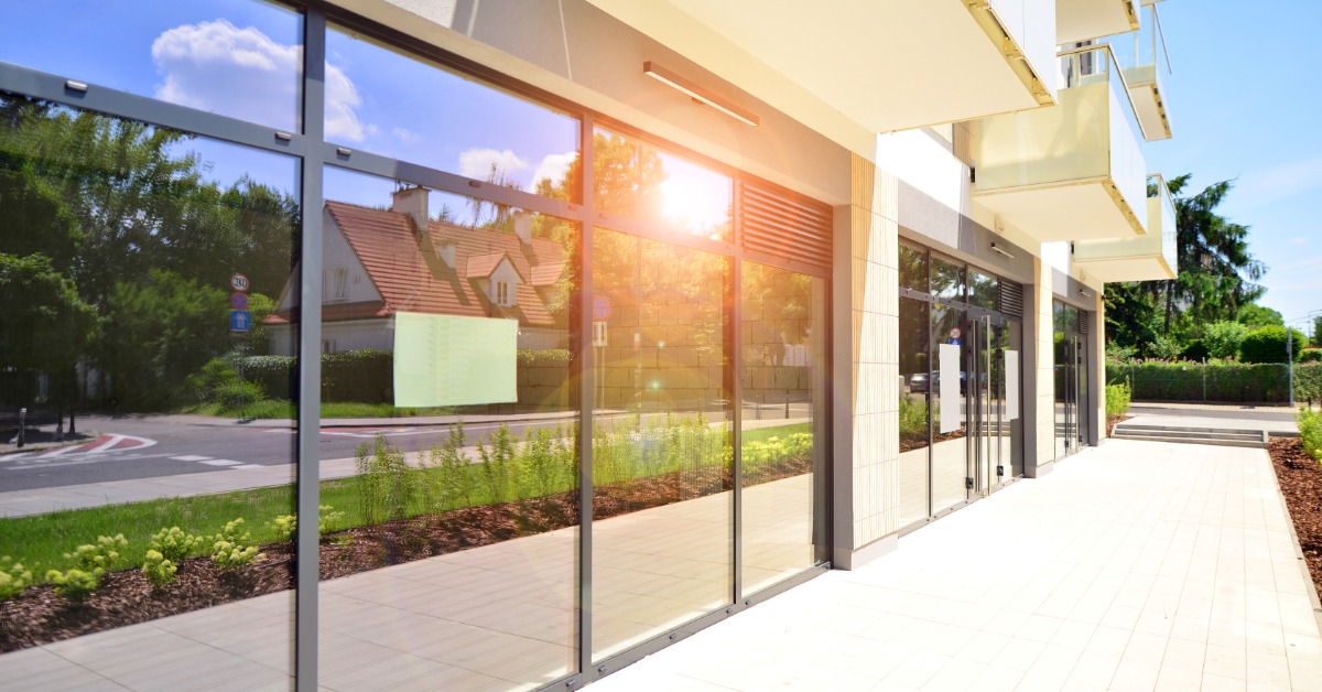A commercial building features large glass storefront windows along a wide sidewalk. Sunlight reflects off the glass.