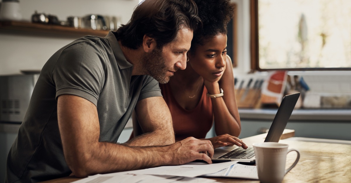 A man and a woman look at a laptop on a wooden table covered with papers. Kitchen shelves and a window are behind them.