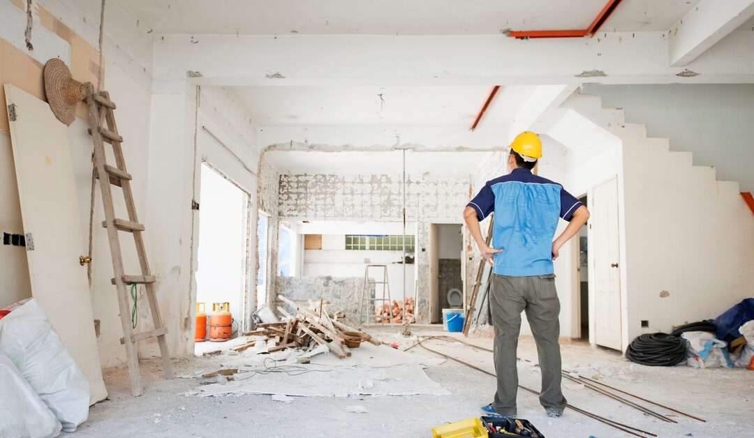 A man wearing a yellow hard hat stands inside a large, partially renovated room filled with construction materials.