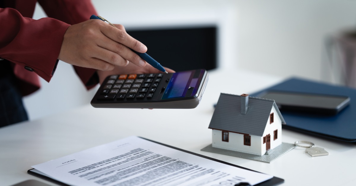 A person points a pen at a calculator screen. They hold the calculator over a miniature house and a clipboard on a desk.