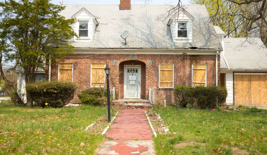 A brick house with boarded-up windows and a damaged roof sits behind a cracked concrete walkway and overgrown lawn.