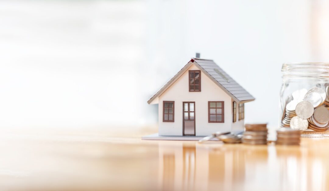 A small model house sits by a glass jar filled with coins. Several stacks of coins are arranged in front of the jar.