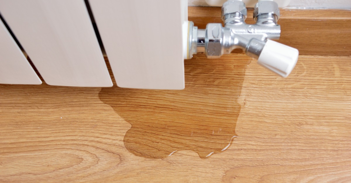 Downward view of a small puddle of water spreading across a wooden floor underneath a metal radiator valve.