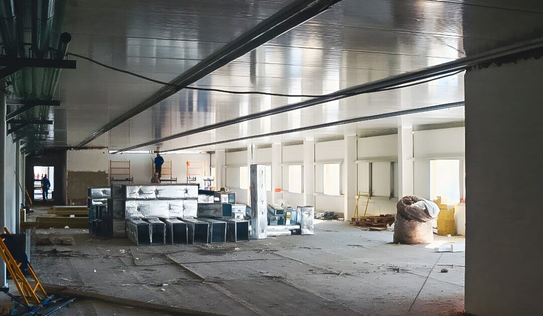 A large commercial space is under renovation. At one end, a worker stands on scaffolding and works near the ceiling.