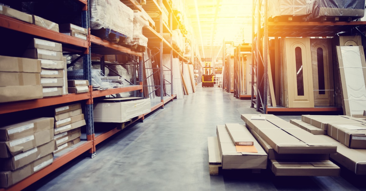 A warehouse aisle is lined with tall metal shelving filled with boxes, doors, panels, and building materials.