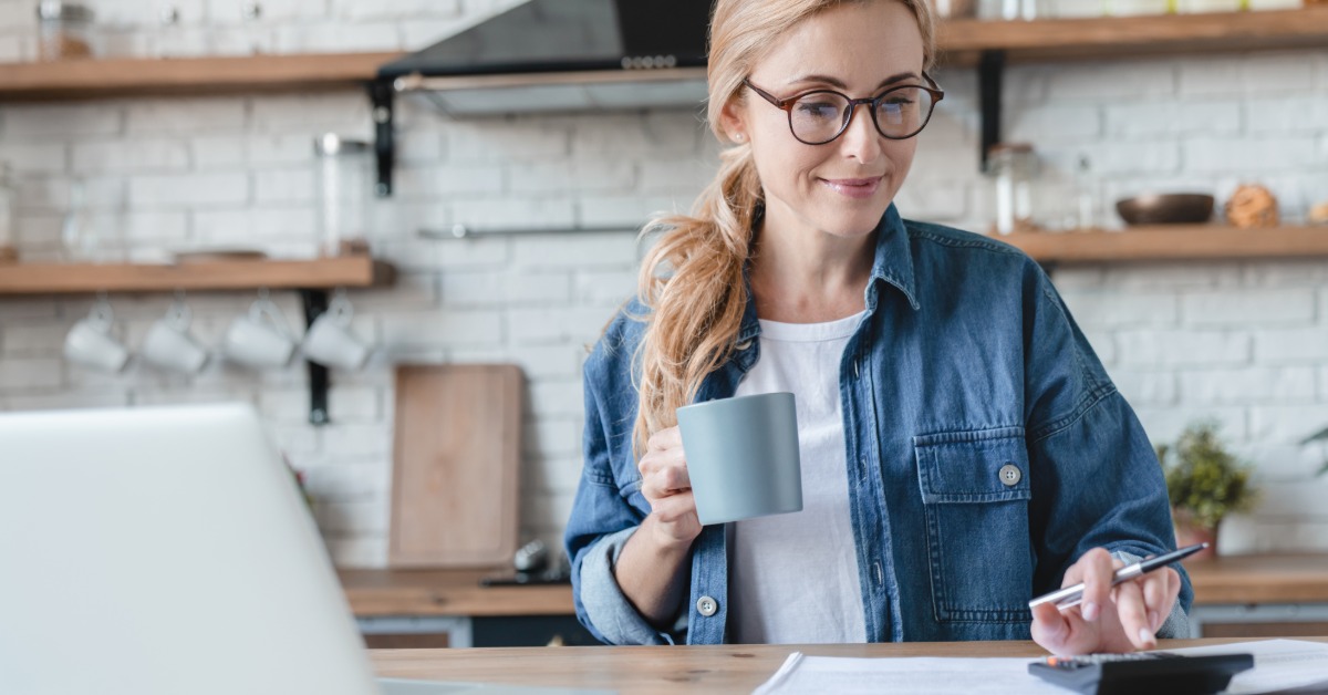 A woman sits at a kitchen table, holding a mug and using a calculator. A laptop and papers are in front of her.