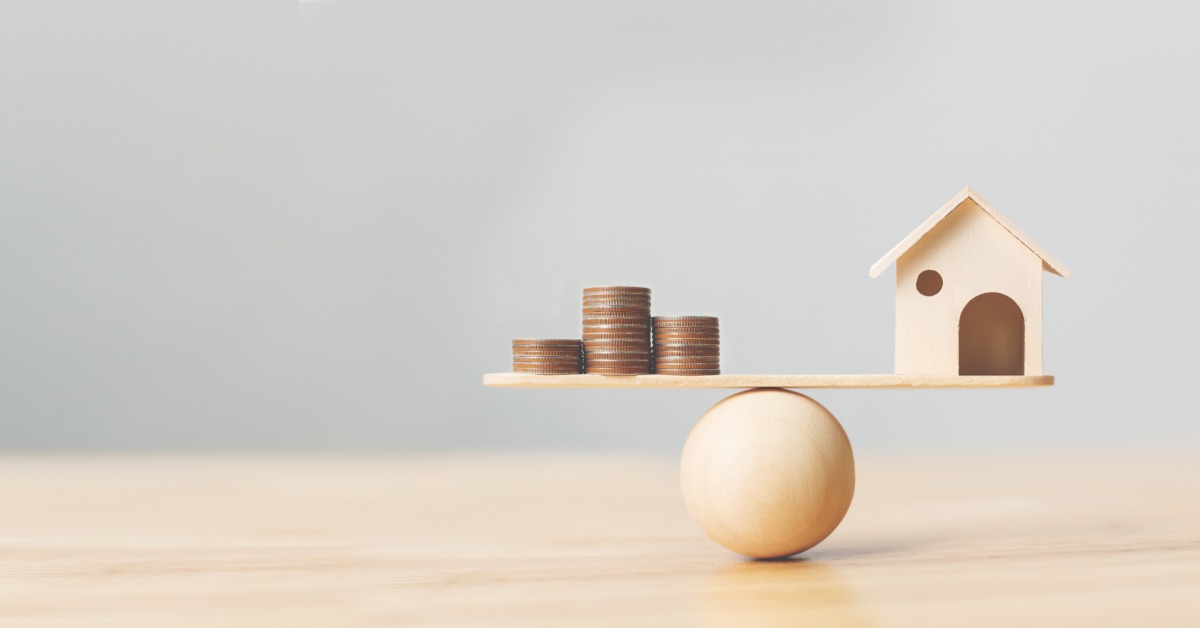 A wooden plank balances on a round wooden sphere. A small house sits on one end, with stacks of coins on the other.