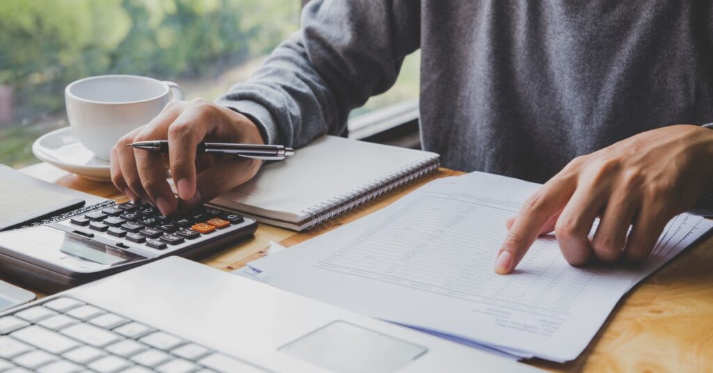 A person uses a calculator and points at lines on a sheet. A laptop, a notebook, and a coffee cup are on the table.