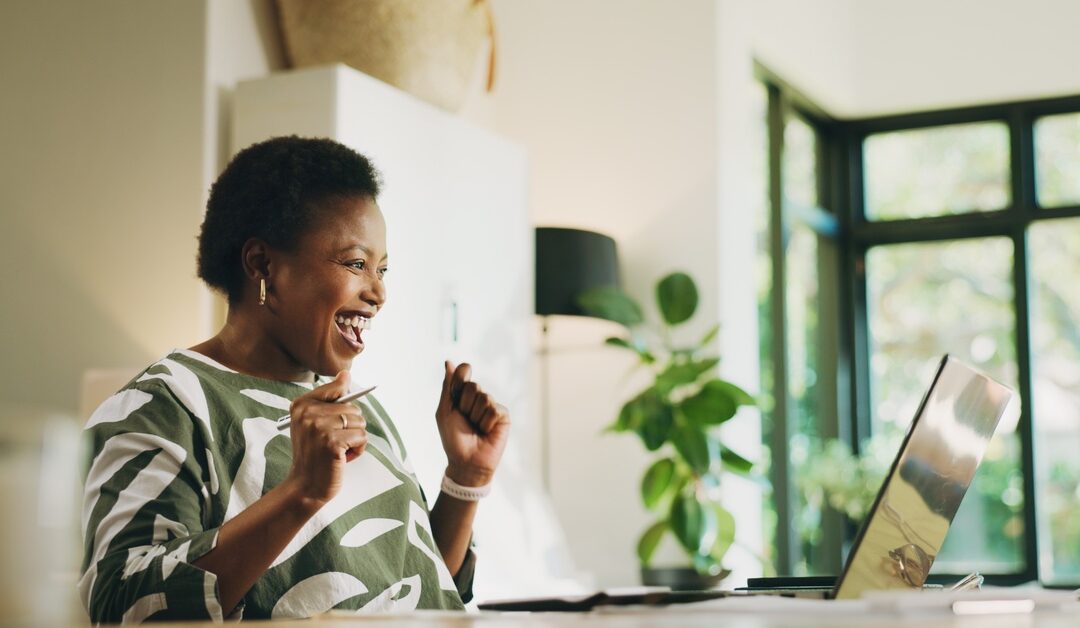 A woman smiles and raises her hands while looking at a laptop. A large window and a potted plant are behind her.