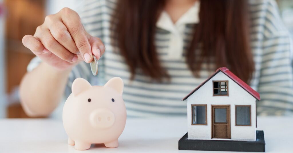 A person places a coin into a pink piggy bank that sits on a table. A small model of a house is next to the piggy bank.