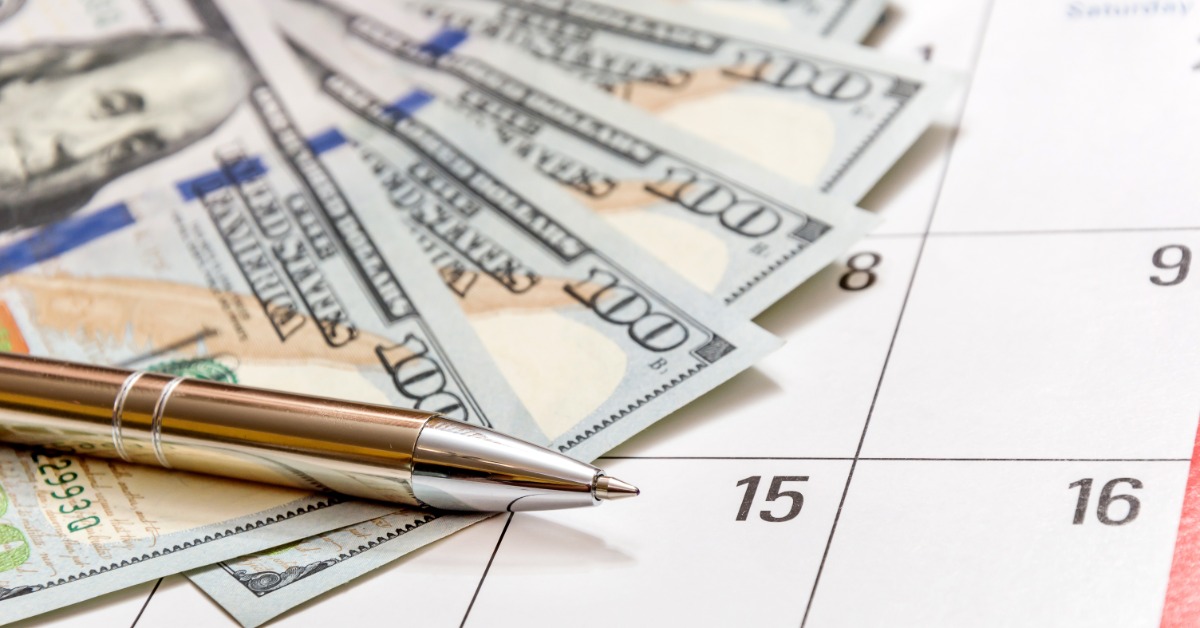 Several crisp U.S. $100 bills are fanned out on top of a paper calendar. A gold-and-silver pen lies on top of the cash.