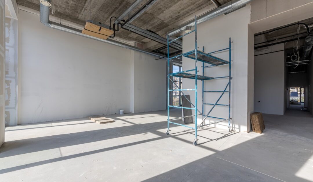 A large, empty space has unfinished white walls and a concrete floor. Blue scaffolding is under exposed ceiling ducts.