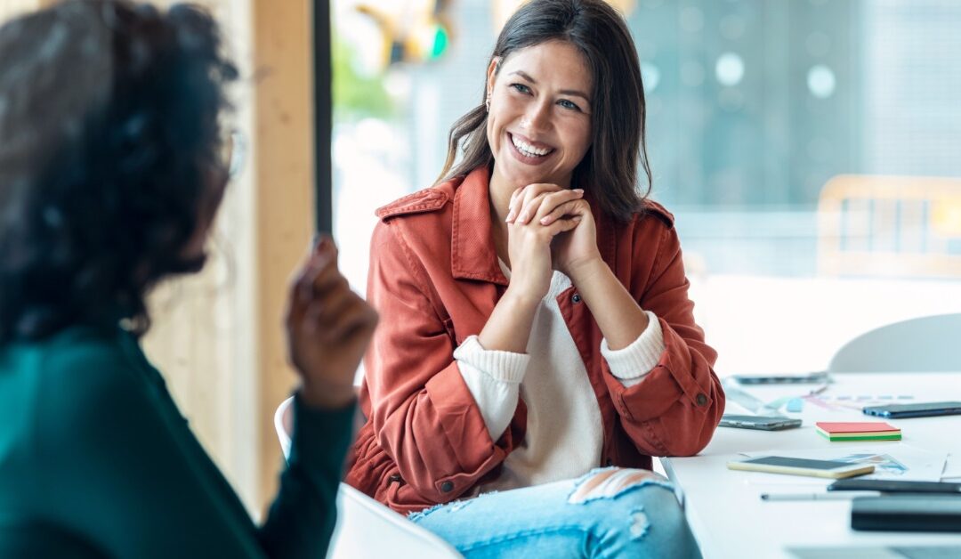 A smiling woman sits at a table with her hands clasped under her chin. She's conversing with a person in the foreground.