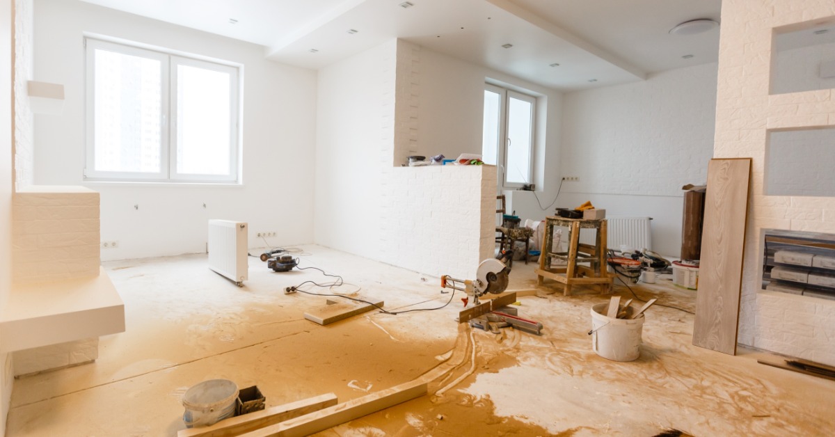 A bright white interior under renovation shows dusty floors, exposed materials, and tools spread throughout the room.