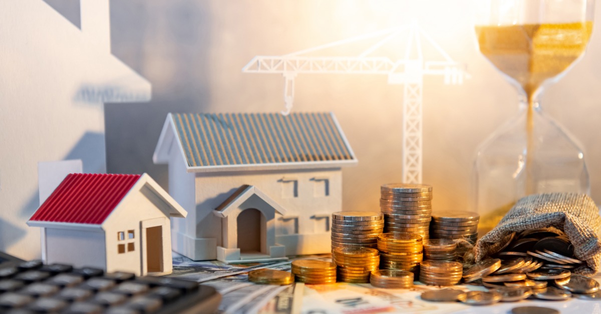 Miniature houses, a sand hourglass, stacks of coins, cash, a calculator, and a miniature crane are on a table.
