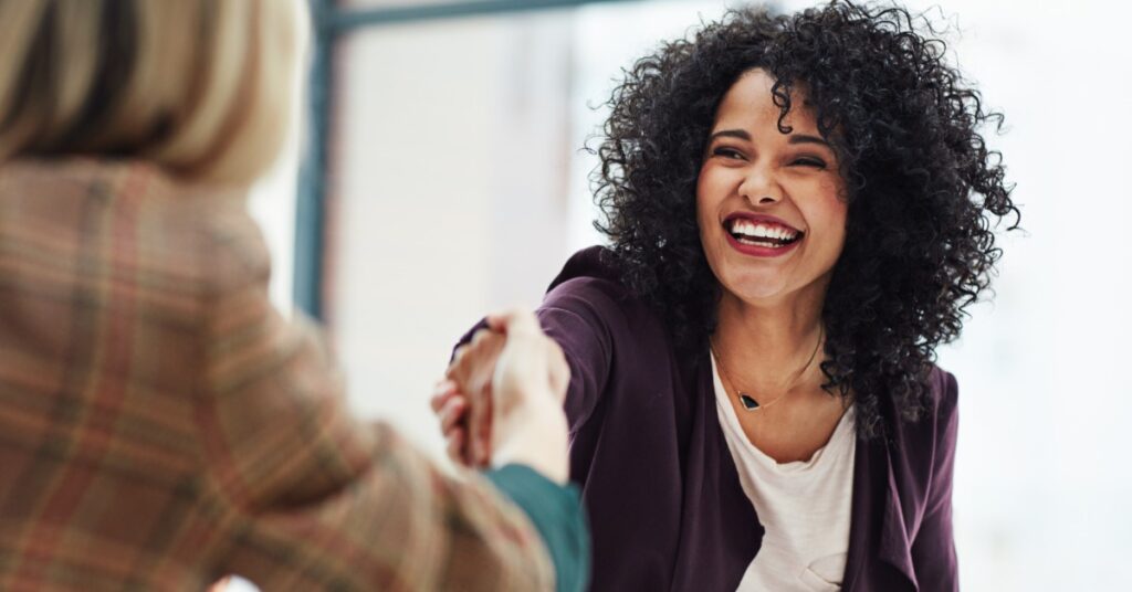 A smiling woman with curly hair shakes hands with a person across the table. The second person is shown from behind.