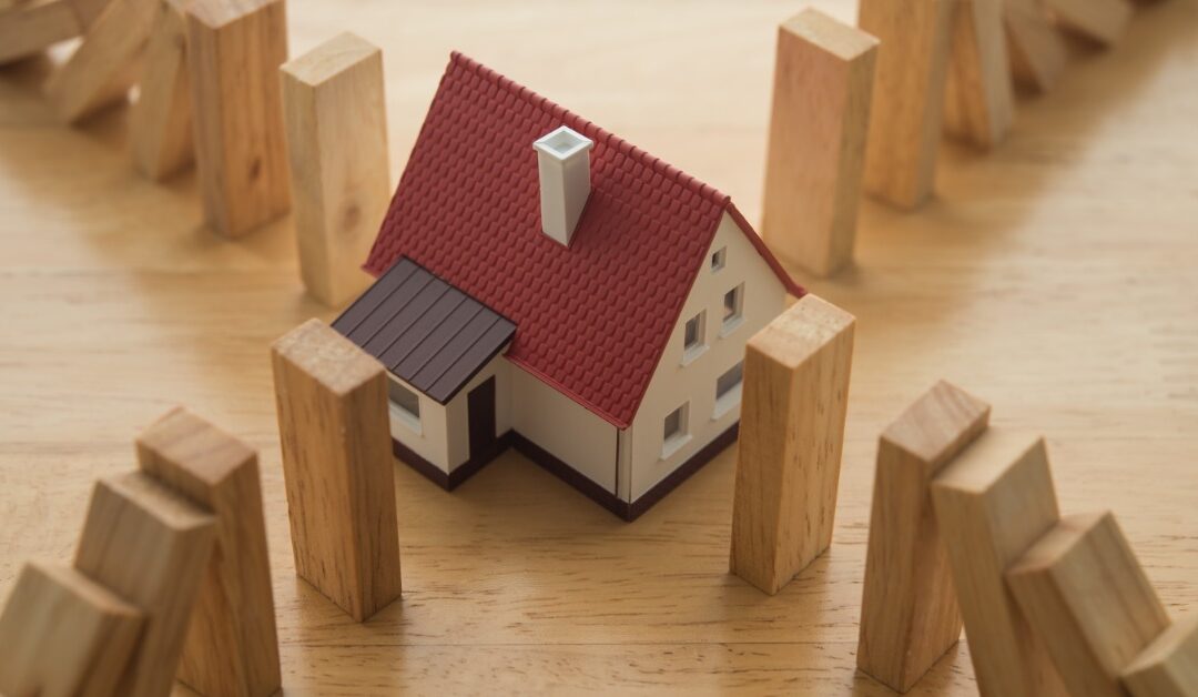 A miniature house is between four radiating lines of wooden blocks. The blocks are tipping over towards the house.