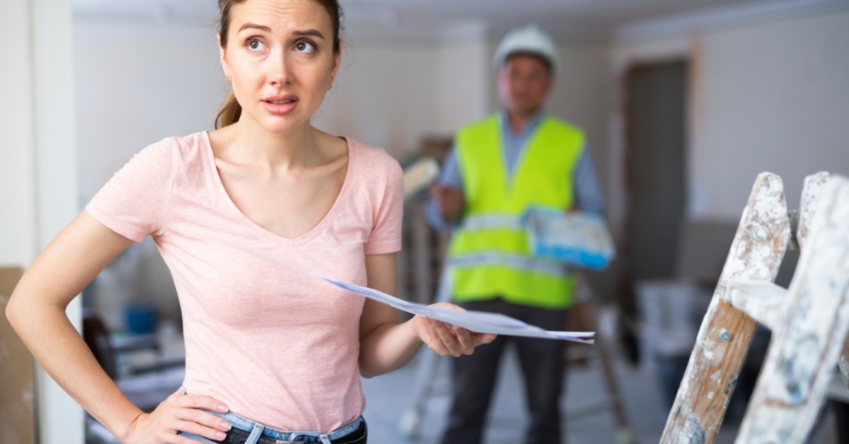 A woman in an unfinished room holds papers and stands near a paint-covered ladder. A worker in a hard hat is behind her.