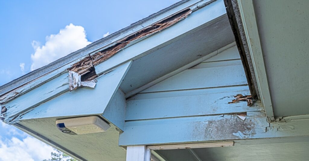A home’s roofline has damaged blue fascia boards, exposed wood, cracked trim, and worn siding by a porch post.