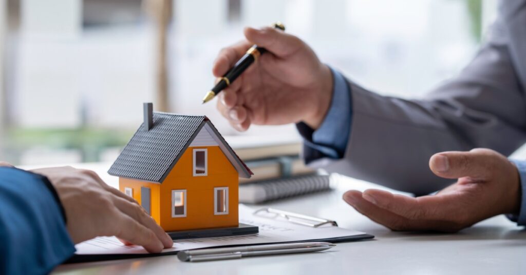 A small orange house model sits atop a clipboard on a table. Two people's hands gesture near the miniature house.