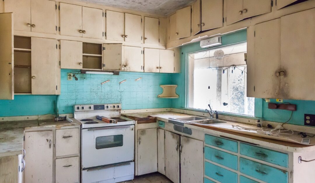 A kitchen has off-white cabinets and turquoise accents. The countertops, cabinets, and floors are stained and damaged.