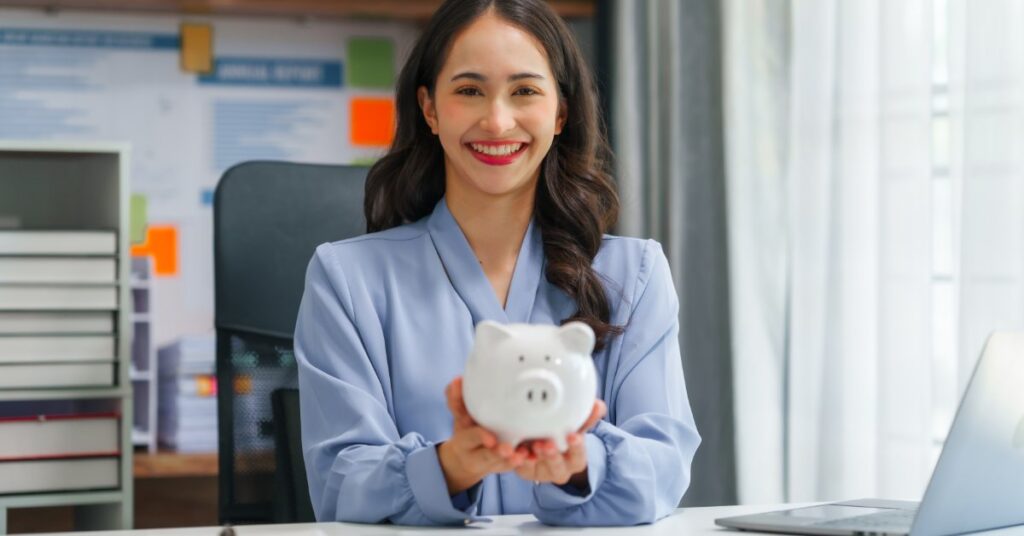 A smiling woman sits in an office, holding a piggy bank. A laptop, a calculator, and a clipboard are in front of her.