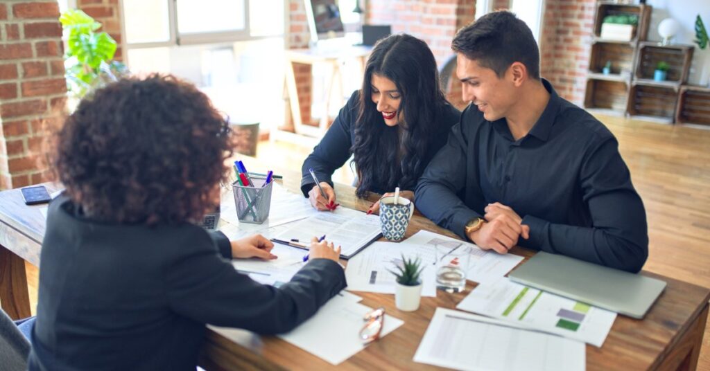 Two people sitting side by side in an office review and sign documents. A person across from them points to one page.