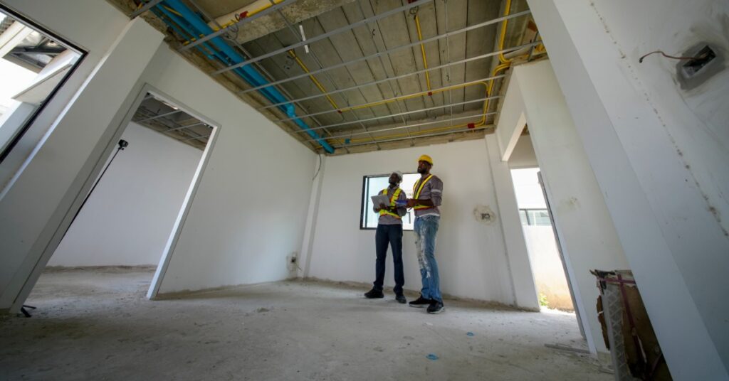 Two construction workers in hard hats stand in an unfinished commercial space with exposed ceiling pipes and framing.