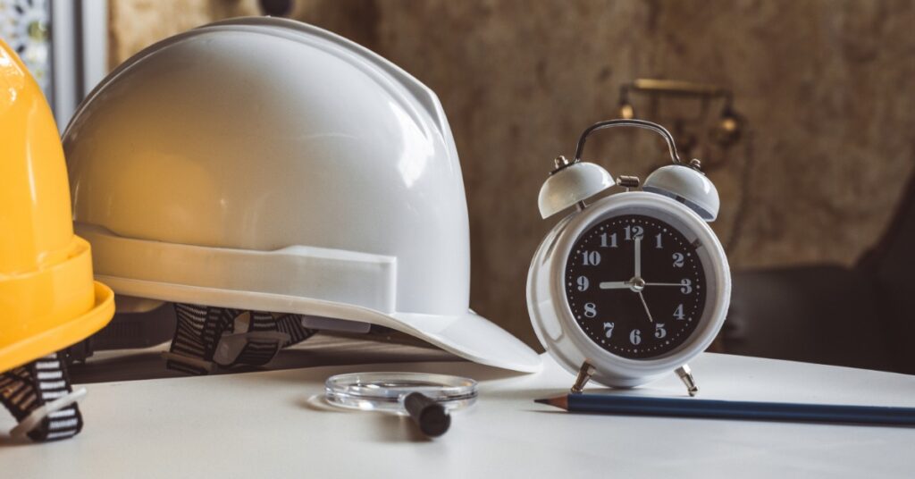 A white hard hat, a yellow hard hat, a black-and-white alarm clock, a pen, and a magnifying glass are on a table.