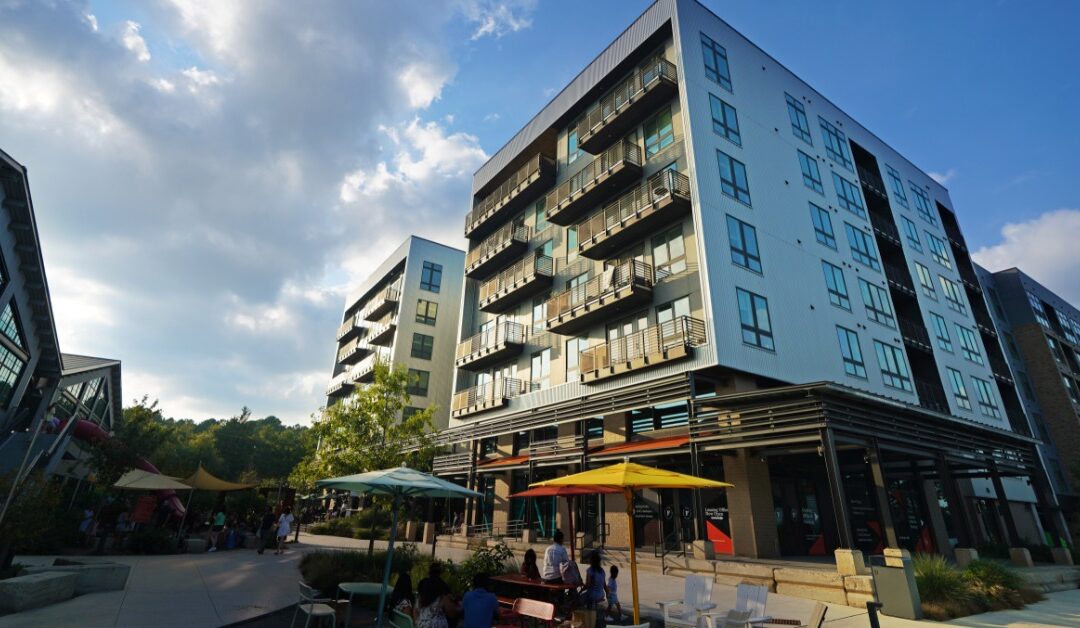 A multi-story building has apartments with balconies. The ground level has storefront-style windows and patio seating.