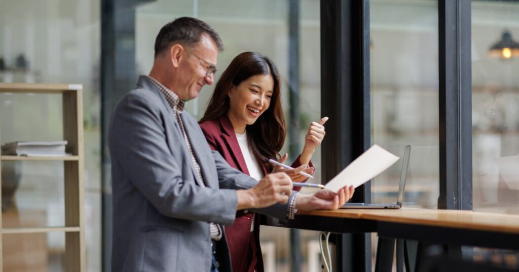 Two people stand at a high table near large windows. One holds a document while the other smiles and gives a thumbs-up.