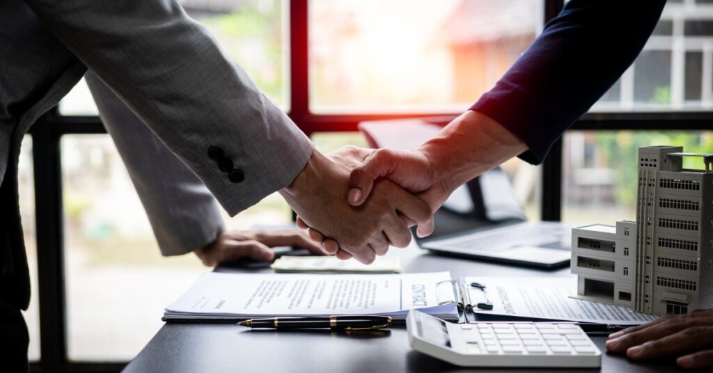 Two people in suits shake hands over a desk. Documents, a calculator, and small building models are on the desk.