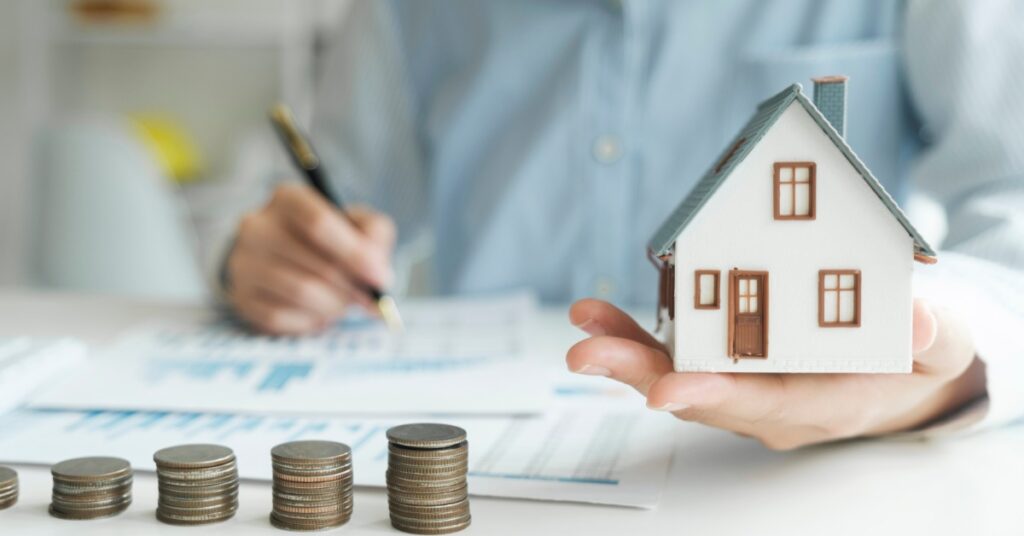 A person holds a small model house in one hand while writing on paperwork with the other. Stacks of coins on the desk.