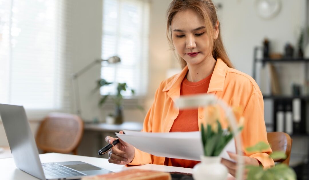A woman sits at a desk with a laptop, reading a document she holds. Books and small plants are on the desk, too.