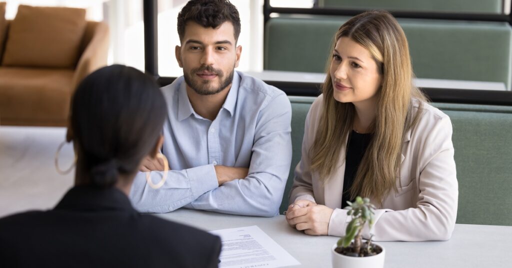 Two people sit across from another person in a cubicle with a low divider wall. A paper is on the table between them.