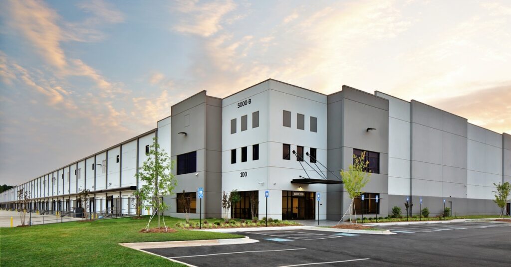 A modern industrial building has a central entrance and loading doors along one side. Young trees are near the entrance.