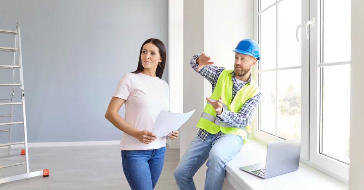 A contractor gestures while sitting on a deep windowsill in an unfurnished room. A woman near him holds documents.