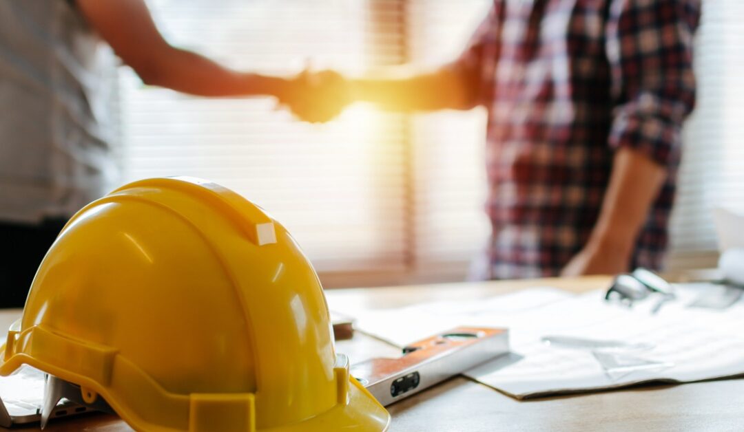 A yellow hard hat is on a wooden table beside papers and a level tool. Two blurred people shake hands in the background.