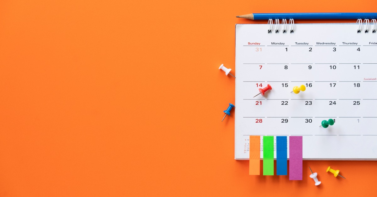 A spiral calendar lies flat and open. Colorful push pins, sticky tabs, and a blue pencil lie on or near the calendar.