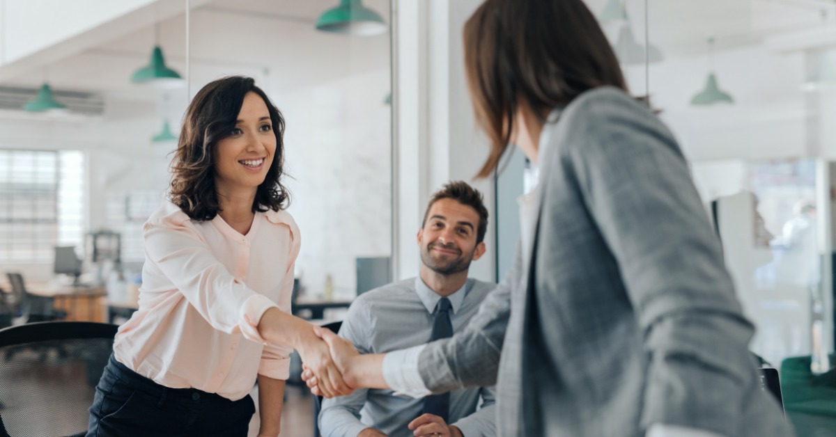 Three people in professional attire are in an office. Two stand and shake hands, while the third sits and smiles.